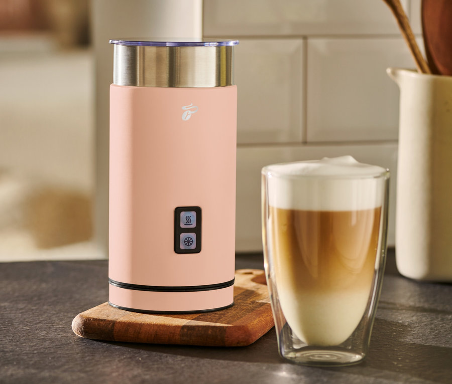 Ein apricotfarbener elektrischer Milchaufschäumer steht neben einem Glas Kaffee.