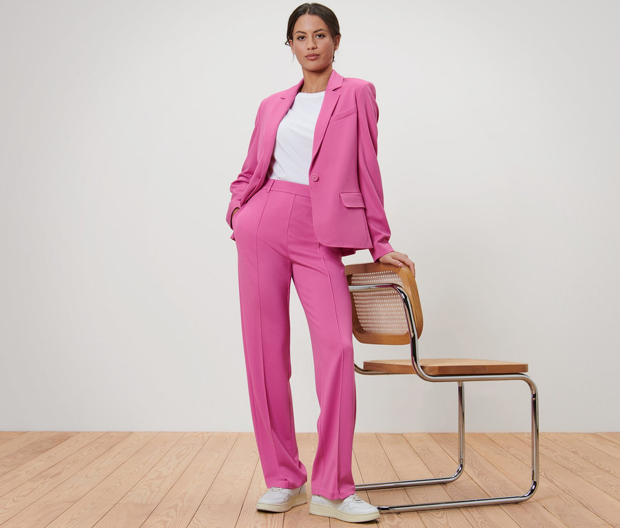 Frau posiert in pinkem Jerseyblazer und Hose neben einem Holzstuhl.