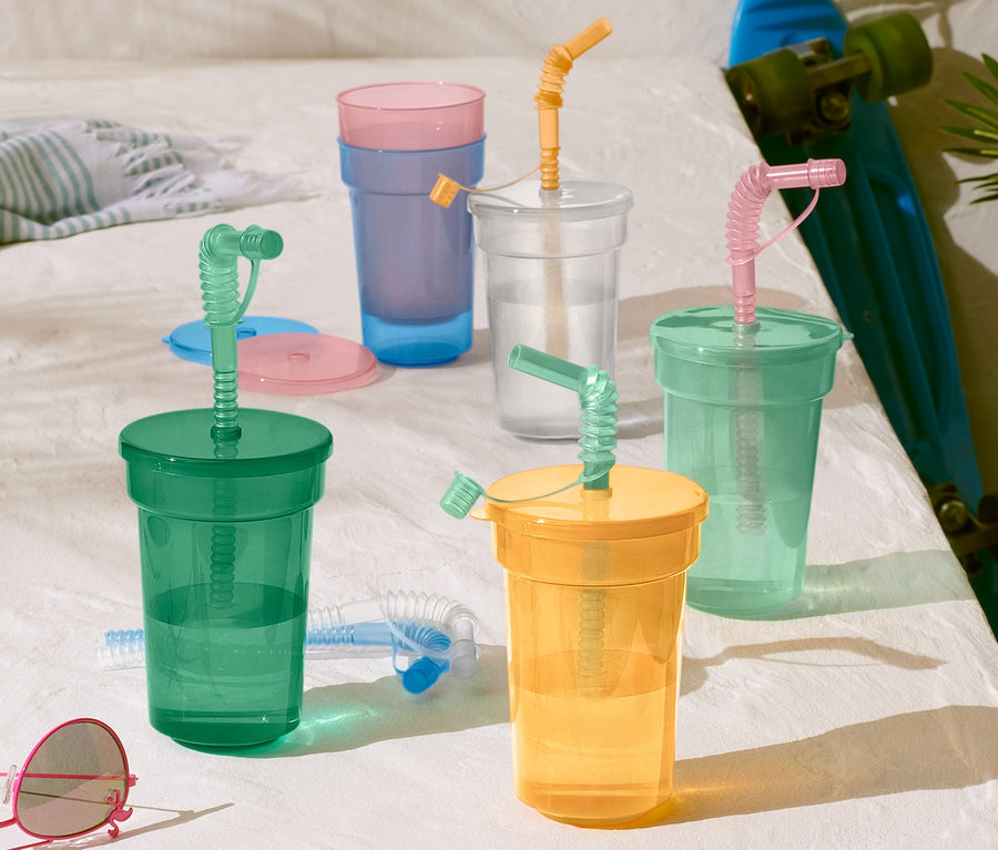 Ein Kinderbecher-Set in Grün, Gelb, Klar und Blau mit Strohhalmen auf einer weißen Oberfläche neben einem Skateboard und einer Sonnenbrille.