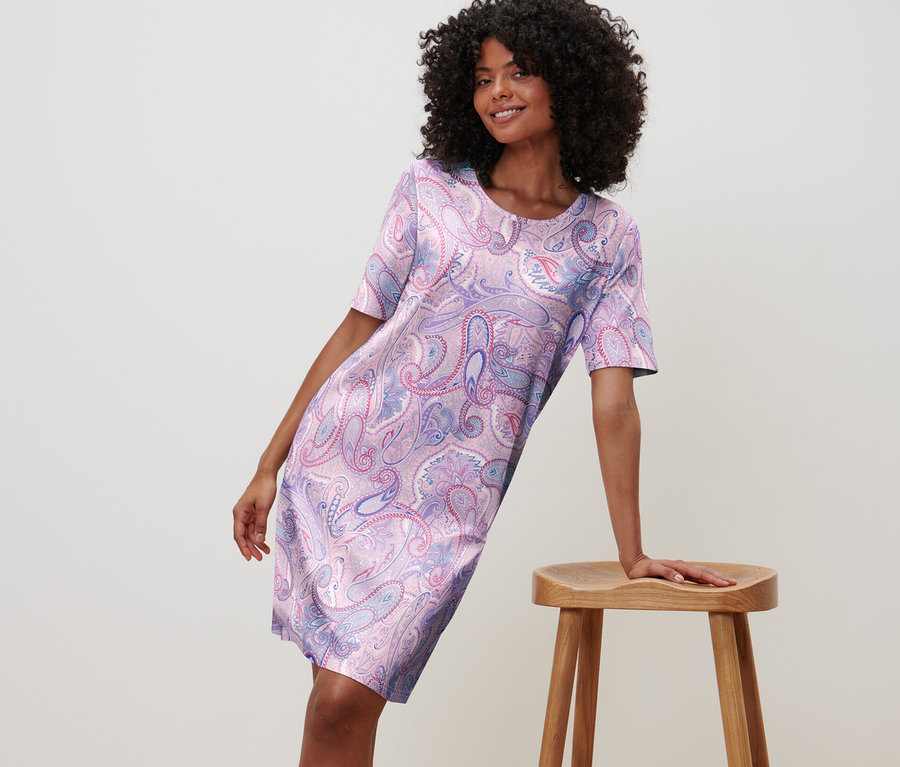 Frau lehnt an Hocker, trägt ein Nachthemd mit Blumenmuster.