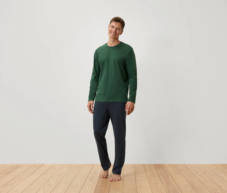 Mann steht barfuß, trägt ein grünes Langarmshirt und dunkelblaue Hosen.