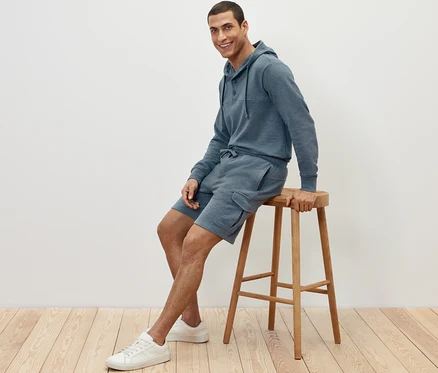 Ein Mann sitzt auf einem Hocker und trägt Cargo-Sweatshorts und einen Hoodie.
