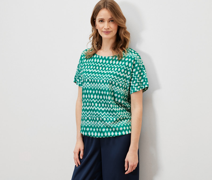 Frau trägt ein bedrucktes Blusenshirt und eine dunkelblaue Hose.