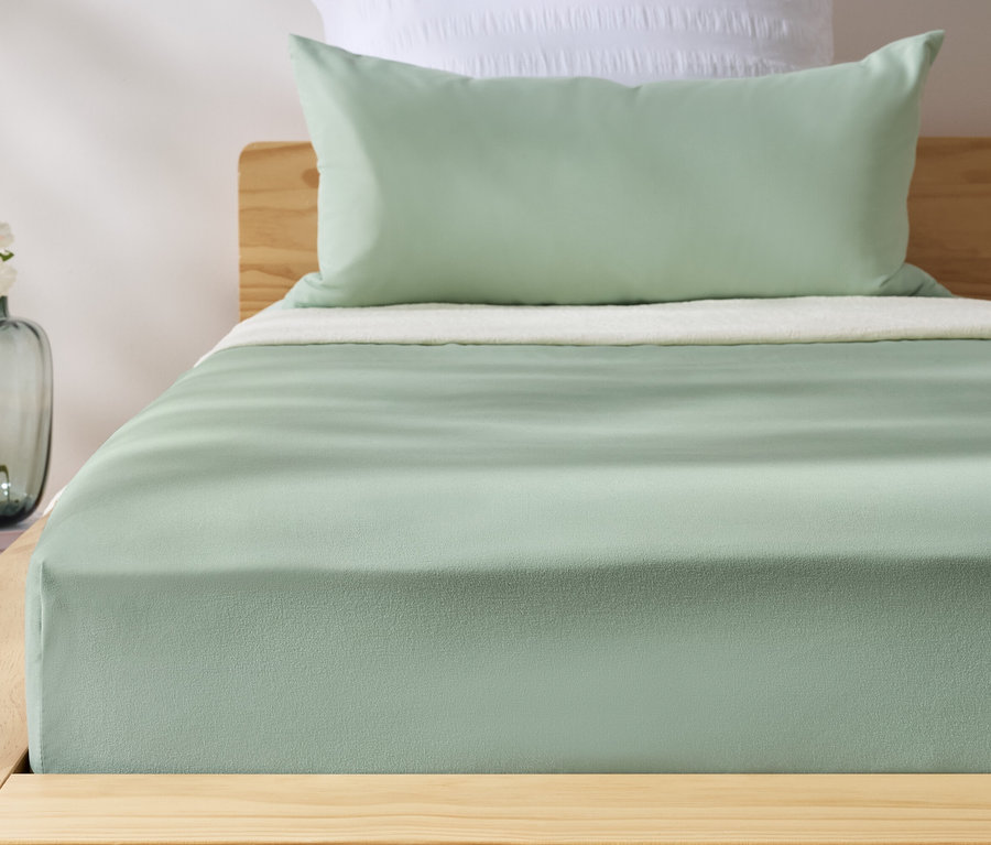 Grünes Microfaser-Spannbettlaken auf einem Bett. Neben dem Bett steht eine Vase mit Blumen.