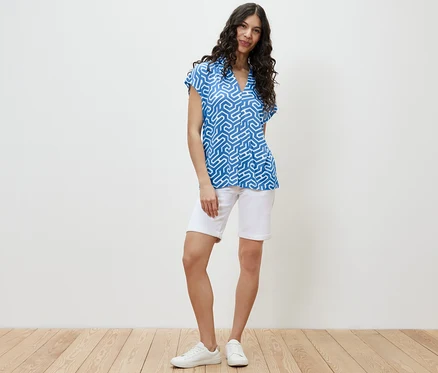 Eine Frau steht und trägt ein blaues Blusenshirt mit V-Ausschnitt und weiße Shorts.