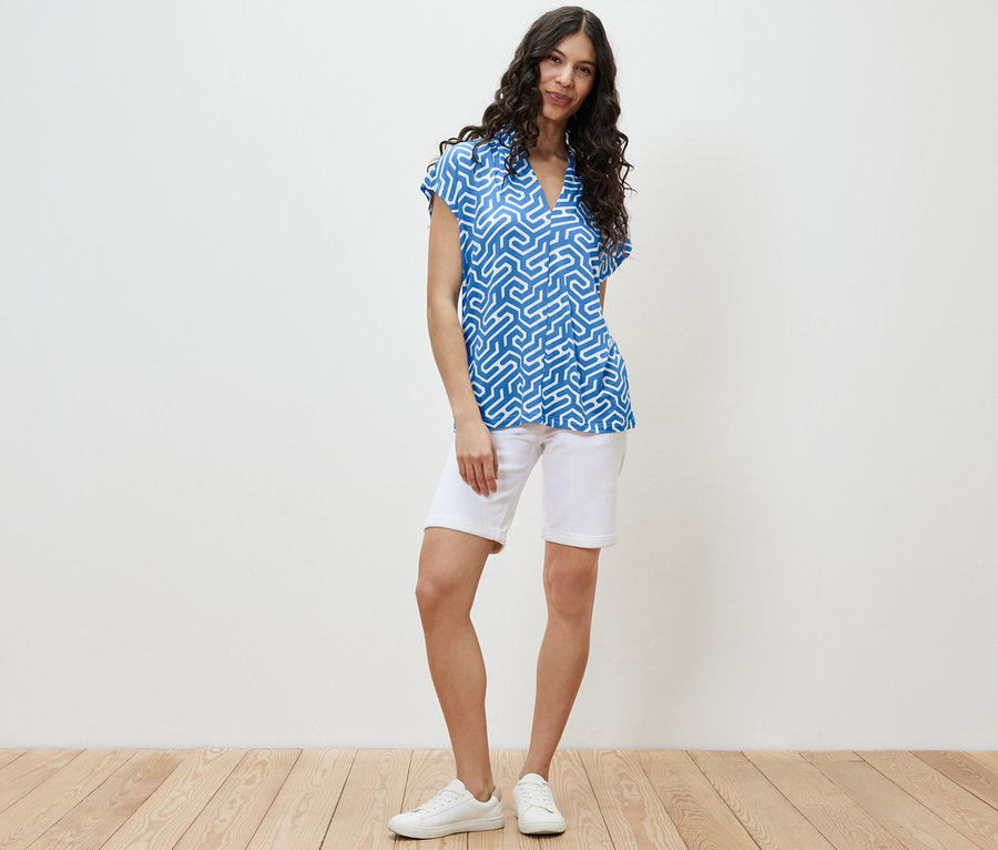 Eine Frau steht und trägt ein blaues Blusenshirt mit V-Ausschnitt und weiße Shorts.