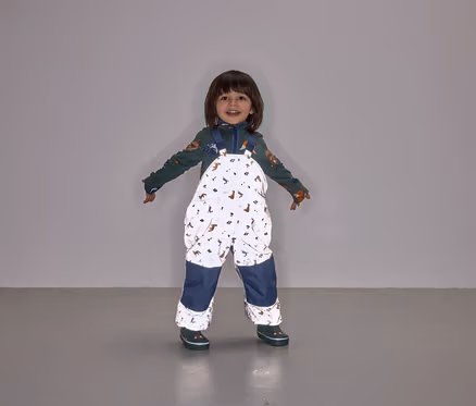 Ein lächelndes Kind steht mit ausgestreckten Armen und trägt eine Latzhose mit Tiermuster und marineblauen Hosenbeinen.