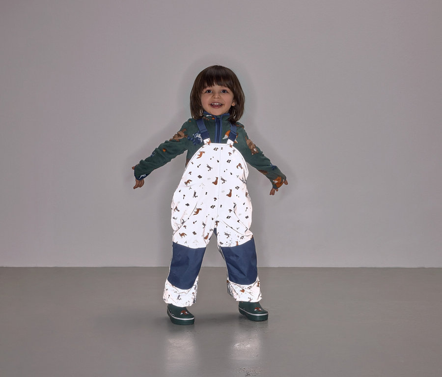 Ein lächelndes Kind steht mit ausgestreckten Armen und trägt eine Latzhose mit Tiermuster und marineblauen Hosenbeinen.