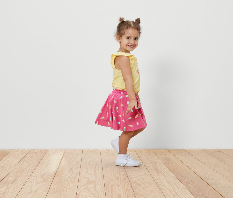 Lächelndes kleines Mädchen posiert seitlich in einem Kinder-Jerseyrock mit Hose mit Eis-Muster und einem gelben T-Shirt.