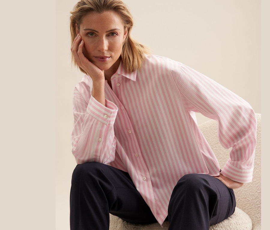 Frau sitzt und posiert in einer hellrosa gestreiften Seidensticker Oversize Hemdbluse, rosé.