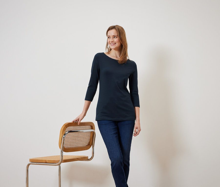 Eine lächelnde Frau in einer marineblauen Bluse mit Dreiviertelärmeln lehnt an einem Holzstuhl.
