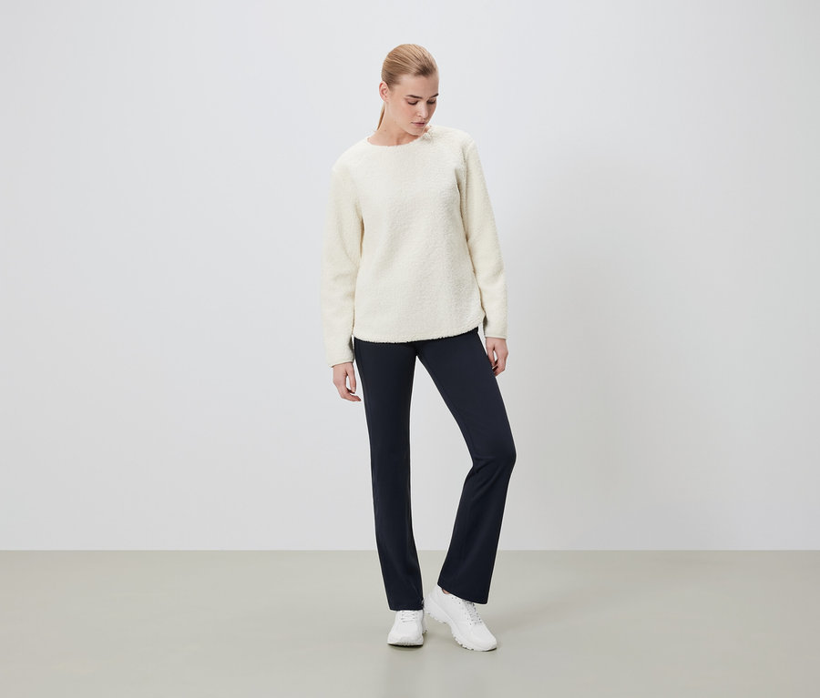 Eine Frau steht und trägt einen cremefarbenen Pullover, dunkelblaue Hosen und weiße Turnschuhe.