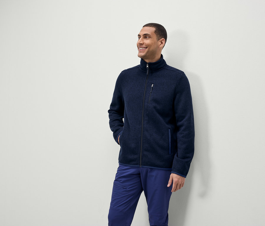 Ein Mann lächelt und schaut zur Seite, er trägt eine blaue Fleecejacke mit Reißverschluss und blaue Hosen.