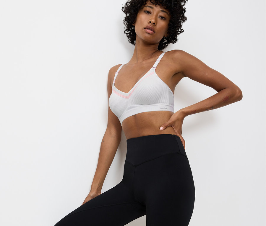 Frau posiert in weißem Sport-BH mit rosa Akzenten und schwarzen Leggings vor einer weißen Wand.