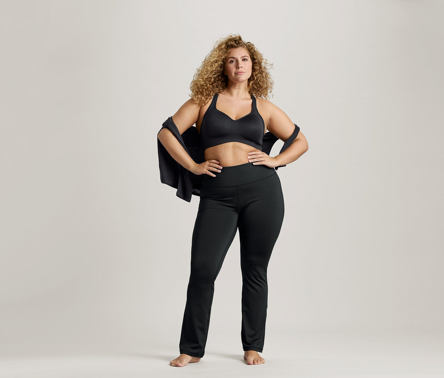 Frau mit lockigem Haar, die einen schwarzen Sport-BH und Leggings trägt, mit einer Jacke über den Schultern und den Händen auf den Hüften.