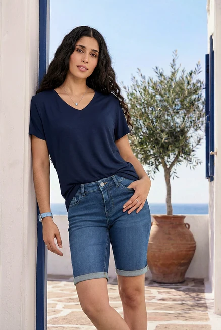 Frau lehnt an Wand, trägt dunkelblaues Shirt, Bermuda-Jeans, blaue und transparente Ringe, Armbanduhr mit Lederband.