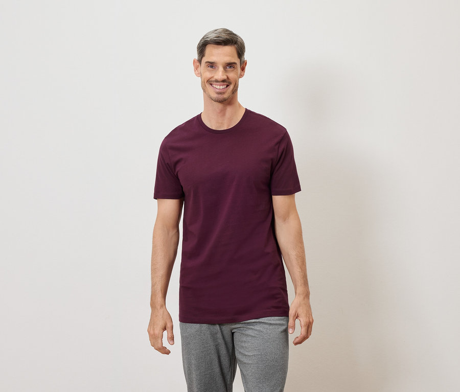 Mann mit grau melierten Haaren lächelt in einem burgunderroten T-Shirt und grauer Hose vor einem weißen Hintergrund.