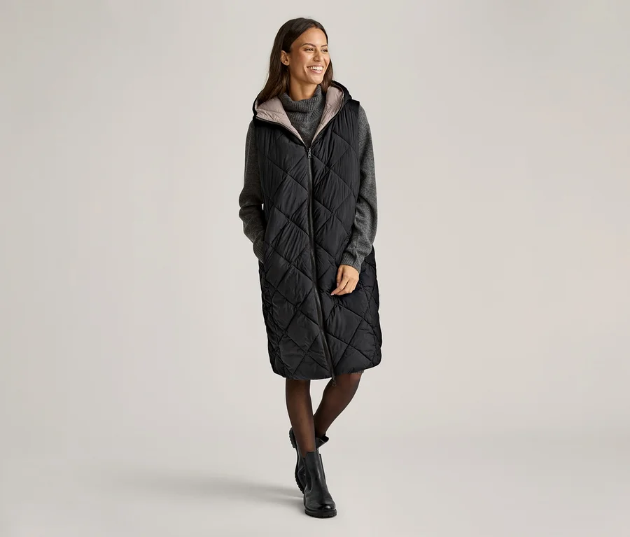 Eine Frau steht und lächelt, sie trägt eine lange, schwarze Steppweste mit Kapuze über einem grauen Pullover, Strumpfhosen und Stiefeln.