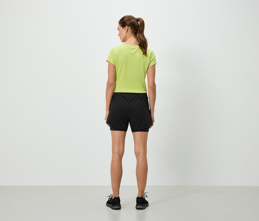 Eine Frau steht mit dem Rücken zur Kamera und trägt ein hellgrünes T-Shirt und schwarze Shorts.