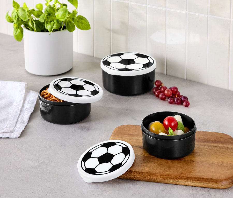 Drei schwarze Vorratsdosen mit Fussballdeckel stehen auf einer Küchentheke, eine Dose ist geöffnet und gefüllt mit Brezeln, eine mit Tomaten und Mozzarella.