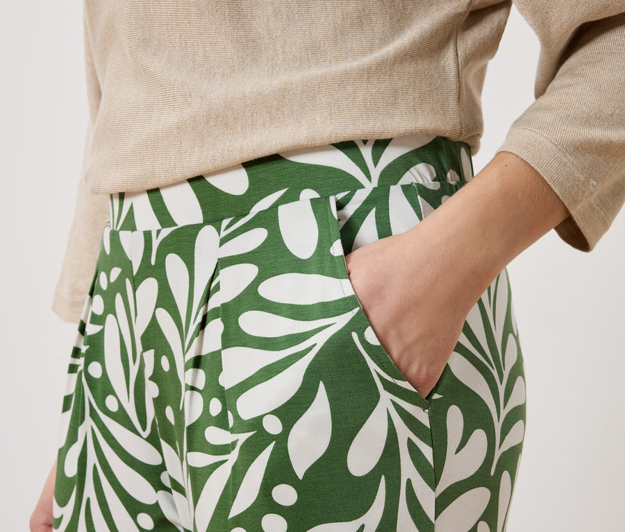 Eine Person trägt eine beige Bluse und eine grüne Hose mit weißem Blätter-Muster. Eine Hand steckt in der Hosentasche.