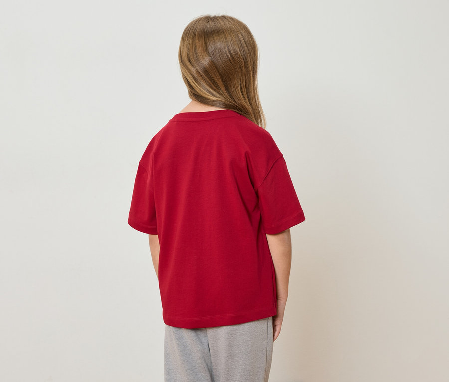 Ein Kind mit blonden Haaren steht mit dem Rücken zur Kamera und trägt ein rotes T-Shirt und graue Hosen.