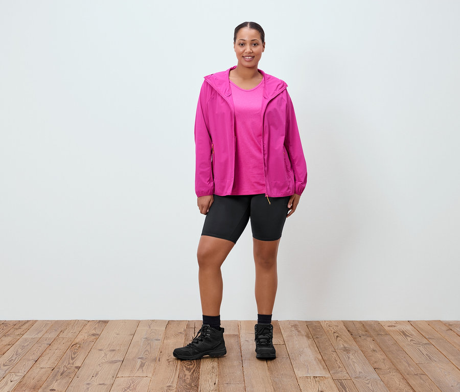 Frau steht und trägt eine pinke Jacke, ein pinkes T-Shirt, kurze schwarze Hosen, schwarze Socken und schwarze Stiefel.