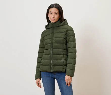 Frau mit olivgrüner Steppjacke mit Kapuze und Reißverschluss, dazu blaue Jeans vor hellem Hintergrund.