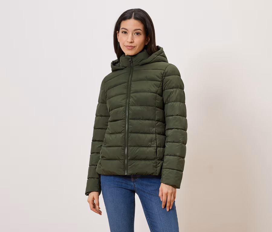 Frau mit olivgrüner Steppjacke mit Kapuze und Reißverschluss, dazu blaue Jeans vor hellem Hintergrund.