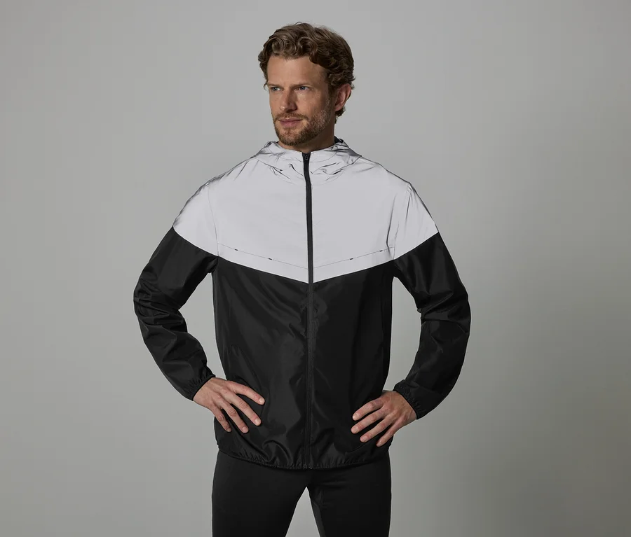 Mann mit Händen auf den Hüften trägt eine schwarz-silberne Windjacke mit Kapuze und schwarze Hose vor grauem Hintergrund.
