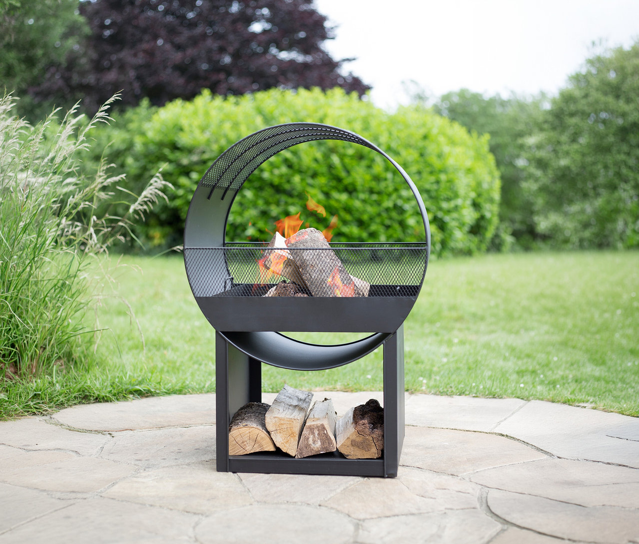 Die Buschbeck Feuerstelle »Porthole« steht im Garten, Holzscheite liegen im unteren Fach und brennen im oberen Bereich.