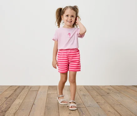 Mädchen mit Zöpfen trägt ein rosa T-Shirt mit einem Seepferdchen-Aufdruck und gestreifte Shorts.