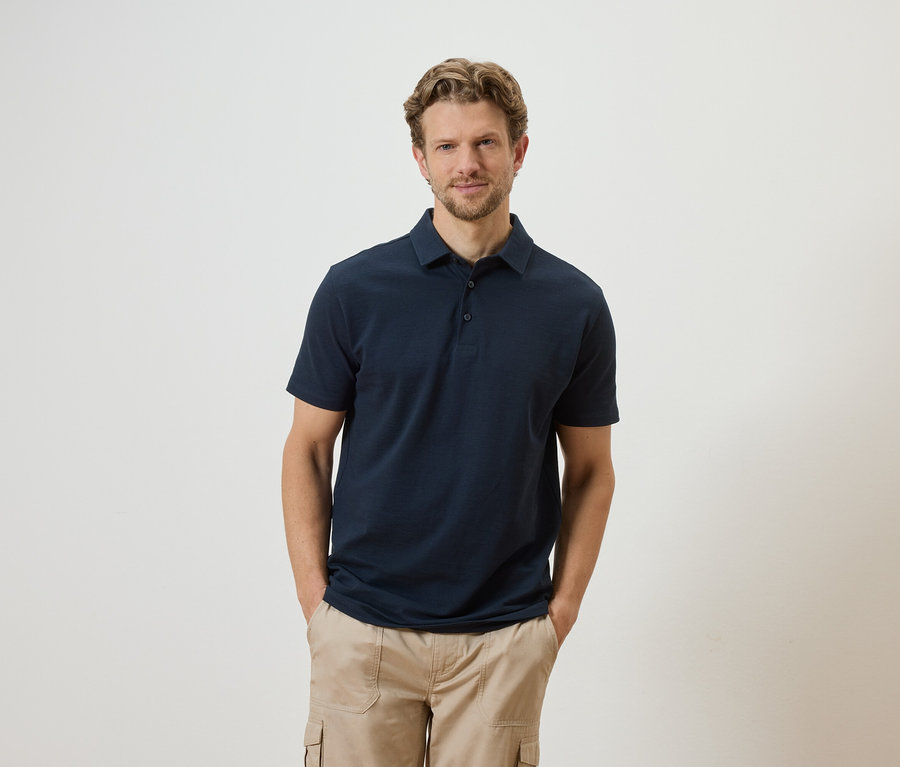 Mann mit blonden Haaren und Bart, bekleidet mit einem dunkelblauen Poloshirt und beigen Cargohosen, posiert vor einem weißen Hintergrund.