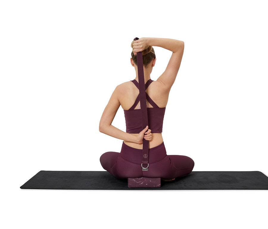 Frau sitzt mit gekreuzten Beinen auf einer Yogamatte und dehnt sich mit einem burgunderfarbenen Yogagurt.