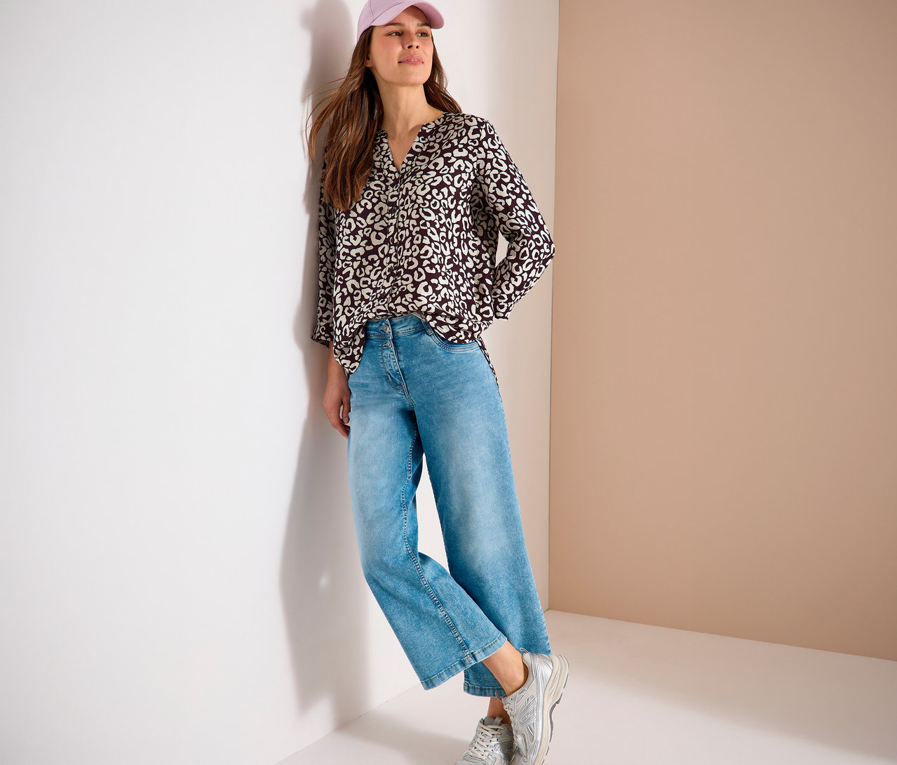 Frau mit rosa Kappe, gemusterter Bluse, blauen Jeans und silbernen Turnschuhen lehnt an einer Wand.