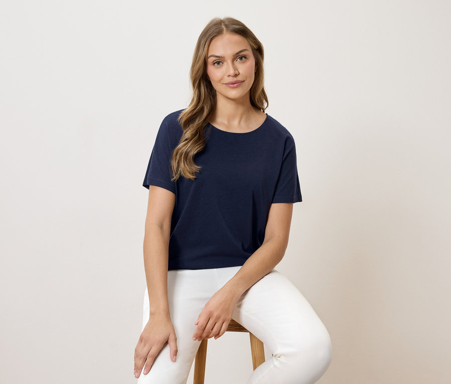 Frau sitzt auf einem Hocker und trägt ein marineblaues T-Shirt und eine weiße Hose.