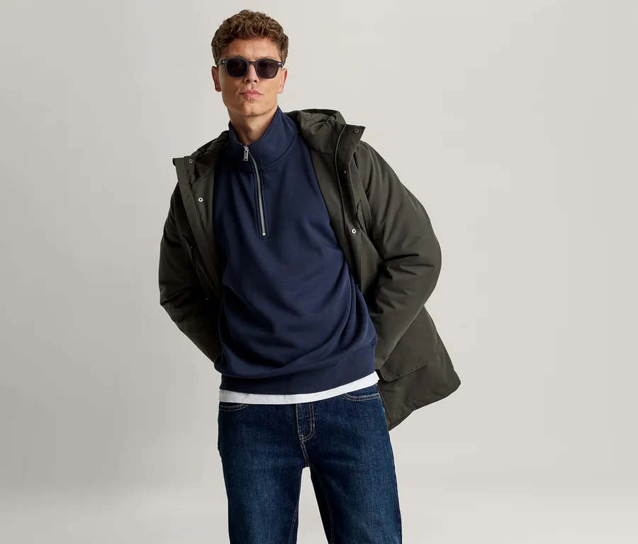 Mann mit Sonnenbrille, dunkelgrünem Kapuzenparka, marineblauem Half-Zip-Sweatshirt, weißem T-Shirt und dunkelblauer Jeans.