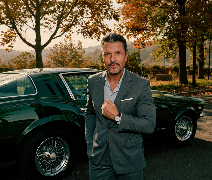 Ein Mann im grauen Anzug und hellblauem Hemd steht vor einem dunkelgrünen Oldtimer. Er hat graues Haar, einen Schnurrbart und einen Ziegenbart und blickt direkt in die Kamera. Am linken Handgelenk trägt er eine silberfarbene Kienzle Uhr »Meister Klassik«. Im Hintergrund sind Herbstbäume und Hügel zu sehen.