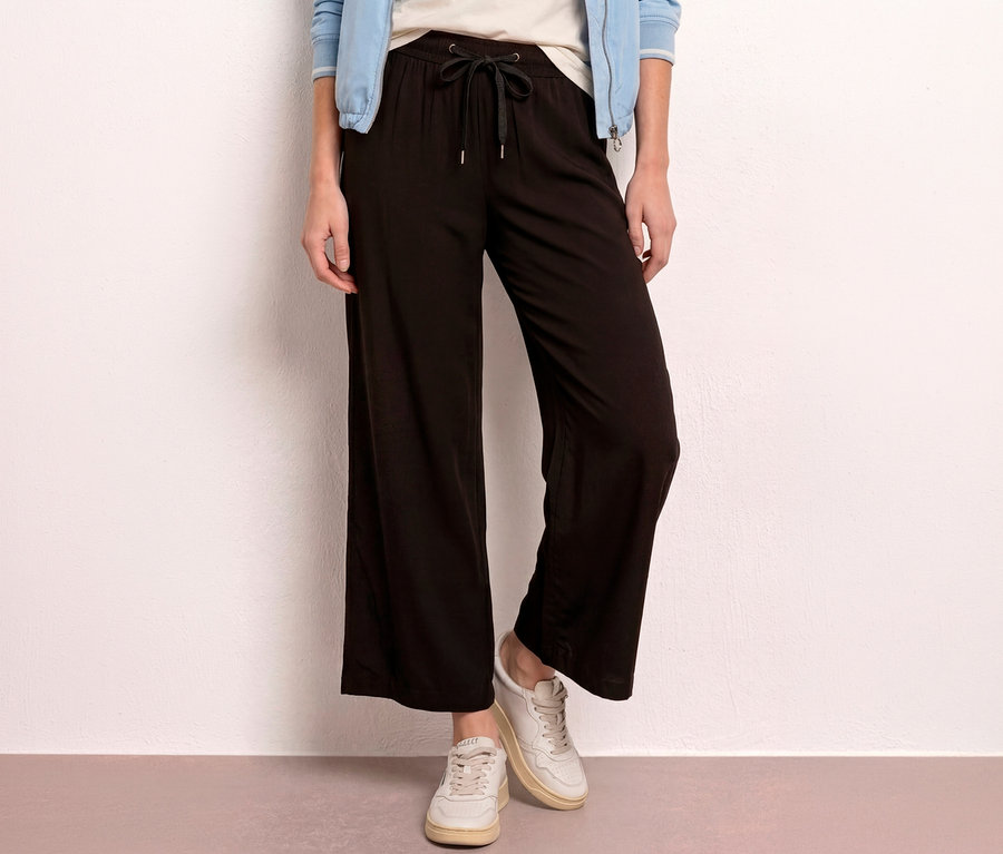 Eine Person trägt eine schwarze Street One 7/8-Culotte »EMMA« mit Kordelzug, ein helles T-Shirt, eine hellblaue Jacke und helle Sneaker.