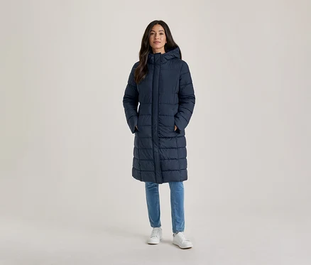 Frau steht, trägt einen langen, marineblauen, wattierten Mantel mit Kapuze, hellblaue Jeans und weiße Turnschuhe, die Hände in den Taschen.