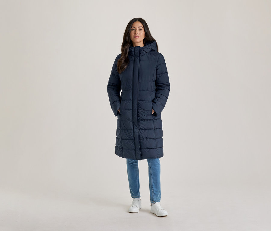 Frau steht, trägt einen langen, marineblauen, wattierten Mantel mit Kapuze, hellblaue Jeans und weiße Turnschuhe, die Hände in den Taschen.