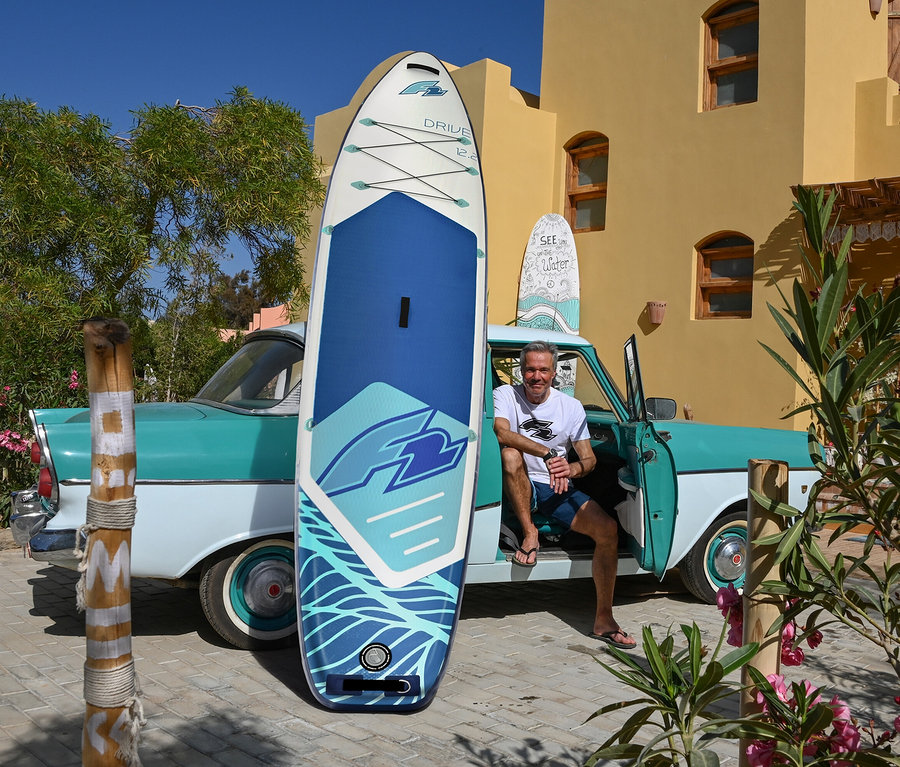 Ein Mann sitzt in einem türkisfarbenen Oldtimer, neben dem ein F2 SUP Board »Drive« aufrecht steht. Im Hintergrund ist ein gelbes Gebäude zu sehen.