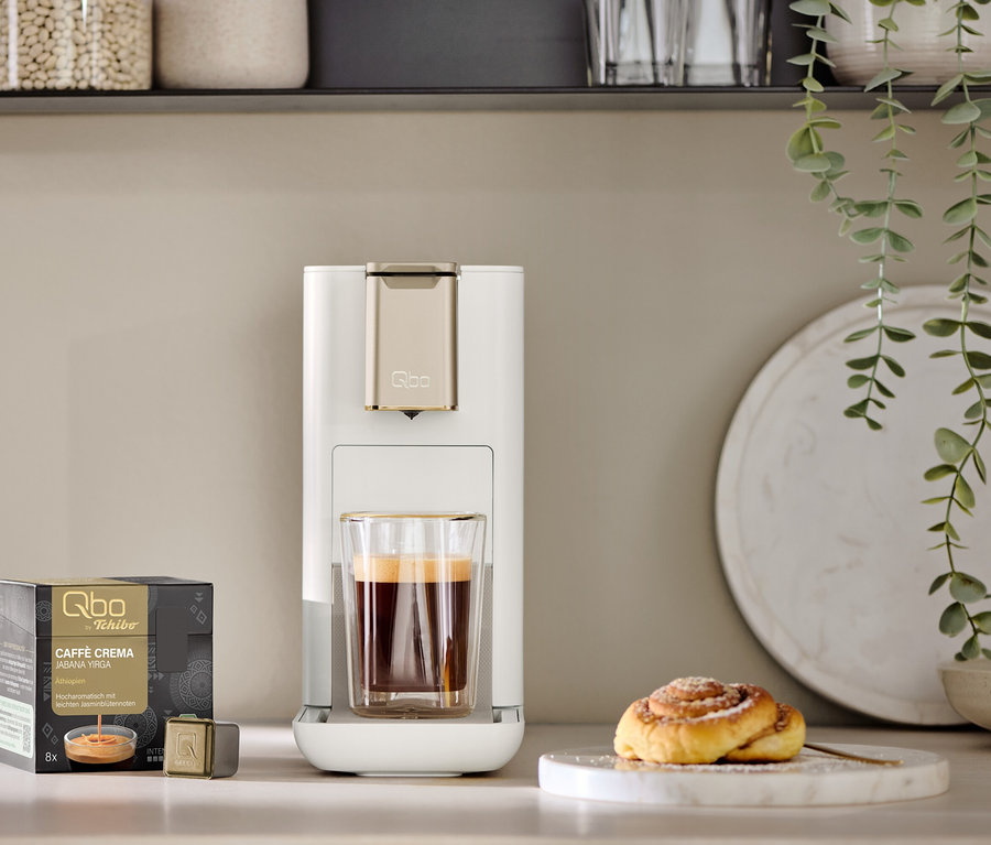 Eine weiße Qbo ICONIC Light Gold Kaffeemaschine mit einer Tasse Kaffee darunter steht auf einer Küchenzeile. Daneben liegen eine Packung Qbo Caffè Crema Jabana Yirga Kapseln und eine Zimtschnecke auf einer Marmorplatte.