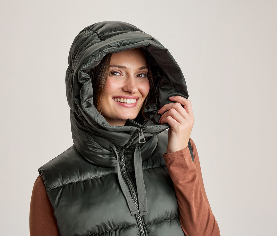 Lächelnde Frau trägt eine dunkelgrüne Steppweste mit großer Kapuze und hält die Kapuze mit der Hand. Darunter trägt sie ein braunes Langarmshirt.