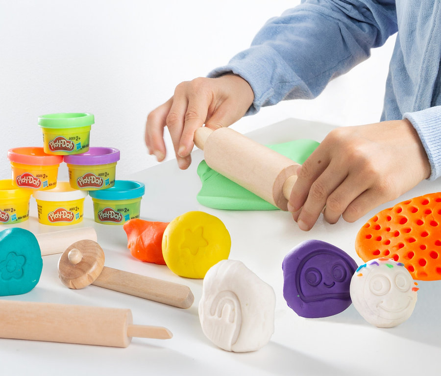 Person rollt grüne Knete mit einer Walze aus. Daneben liegt das roba x Play-Doh Sitzgruppen-Set und weitere Knetfiguren.