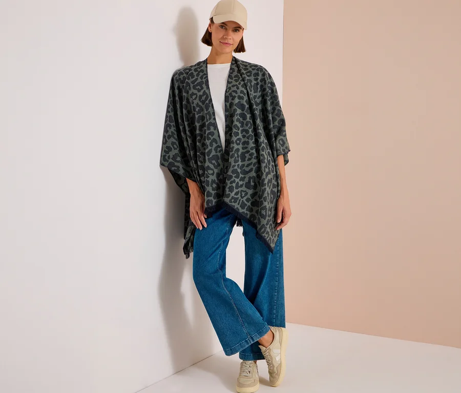 Frau trägt beigefarbene Kappe, weißes T-Shirt, blaue Jeans, beigefarbene Sneaker und einen dunkelgrauen CECIL Poncho mit Leopardenmuster, an eine Wand gelehnt.