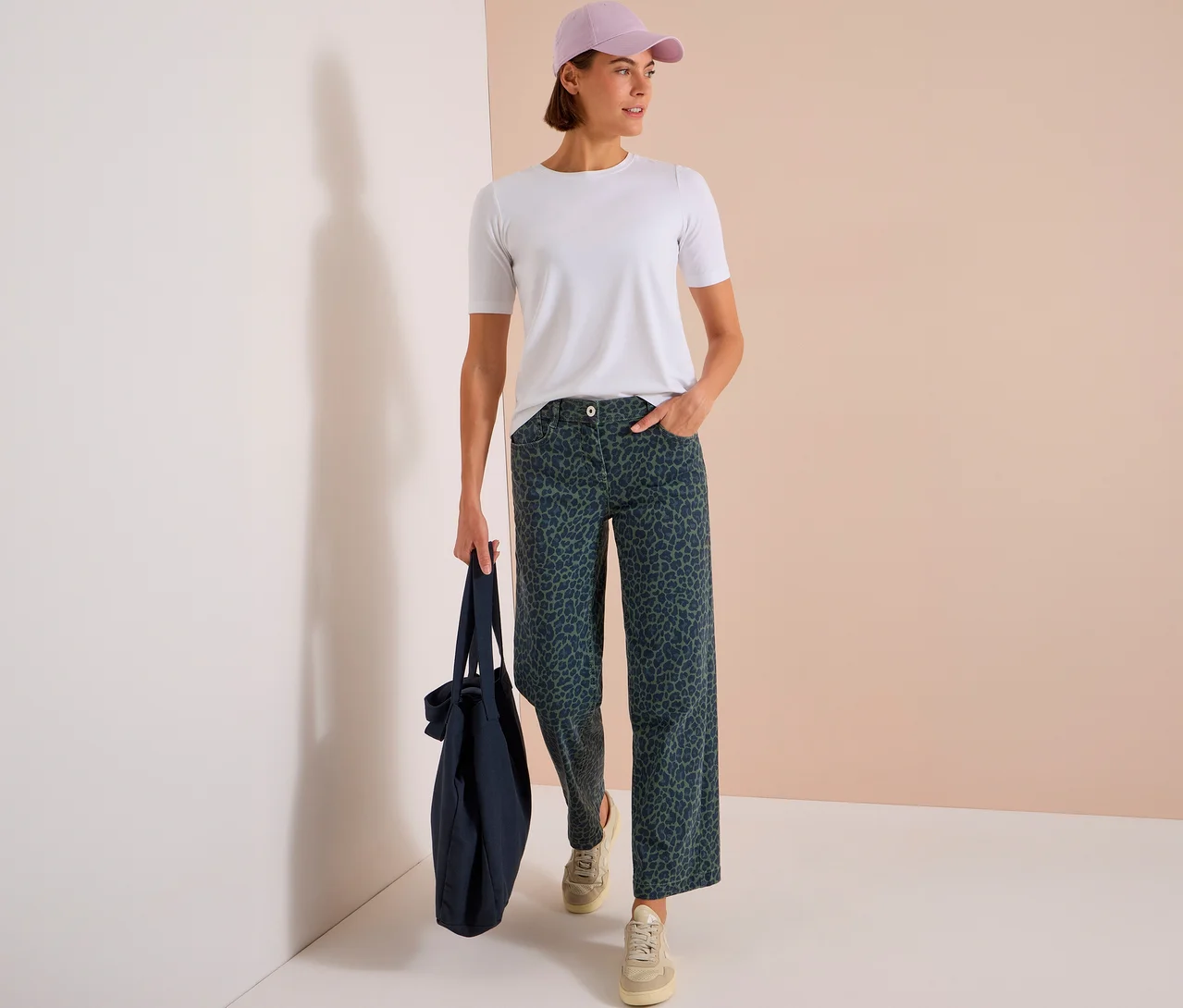 Stehende Frau mit weißem T-Shirt, hellvioletter Kappe und dunkelgrüner CECIL Jeans mit Leopardenmuster, die eine dunkelblaue Einkaufstasche hält.
