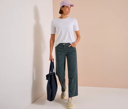 Stehende Frau mit weißem T-Shirt, hellvioletter Kappe und dunkelgrüner CECIL Jeans mit Leopardenmuster, die eine dunkelblaue Einkaufstasche hält.