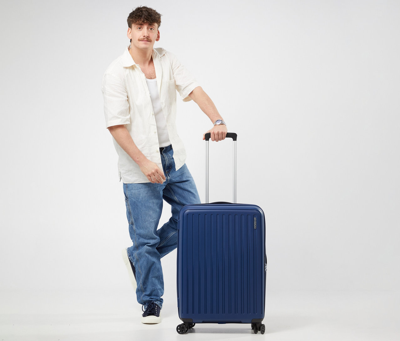 Ein Mann steht mit einem Fuß auf einem mittelgroßen American Tourister Rejoy Spinner Koffer in Navy Blue und hält dessen Griff.