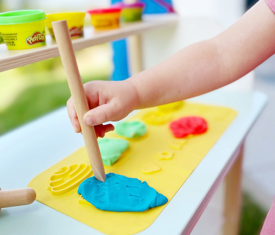 Ein Kind formt blaue Knete mit einem Holzwerkzeug auf einer gelben Matte des roba x Play-Doh Sitzgruppen-Sets.